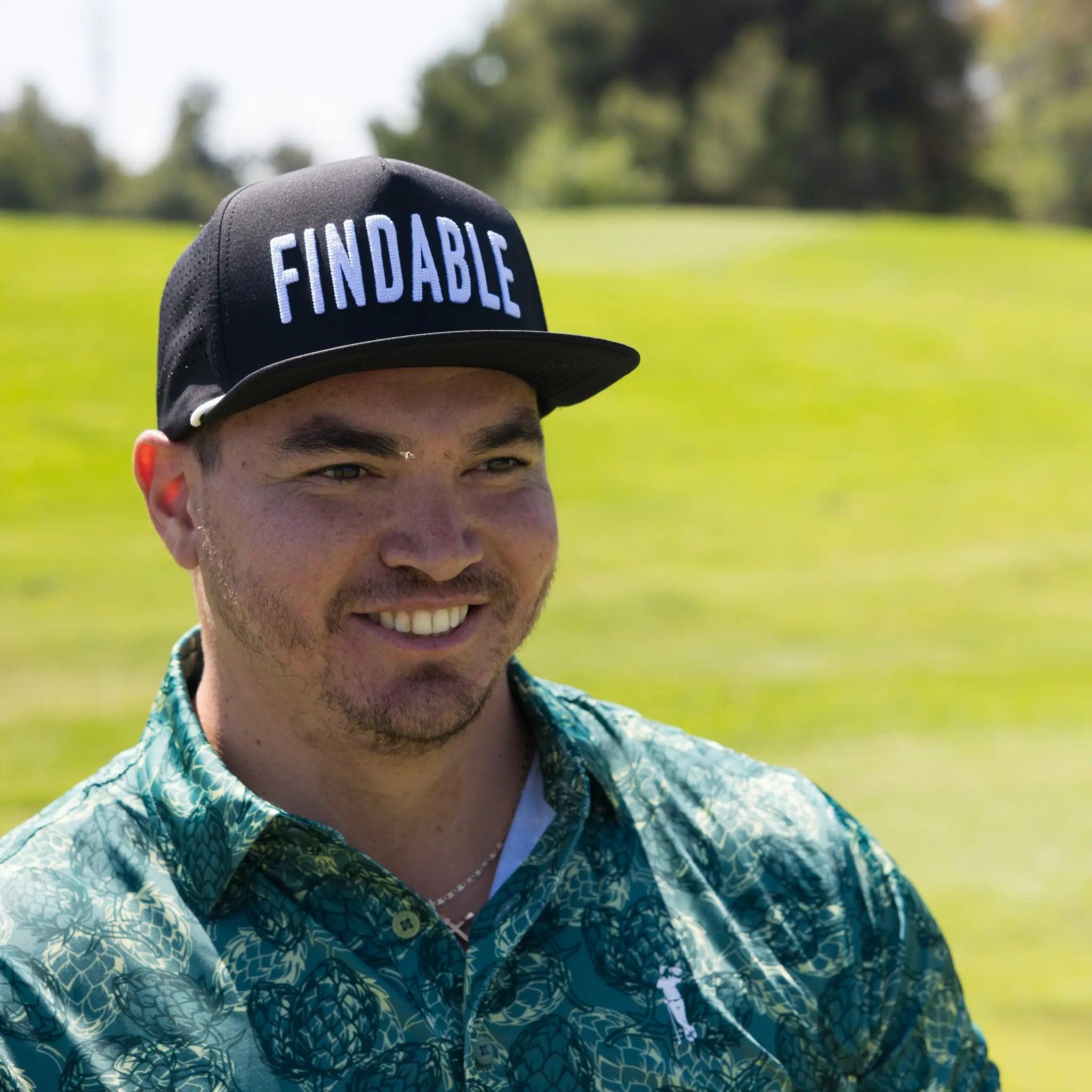 A man wearing the Grunk Dolfer Findable Hat—a black curved brim golf cap with "FINDABLE" on the front—stands outdoors in a green patterned shirt, with a grassy field and trees behind him on a sunny day.