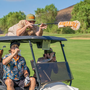 Three men in a golf cart on a golf course; one aims a large paddle labeled FINDABLE, while the others drink and look through binoculars. All wear casual shirts and hats.