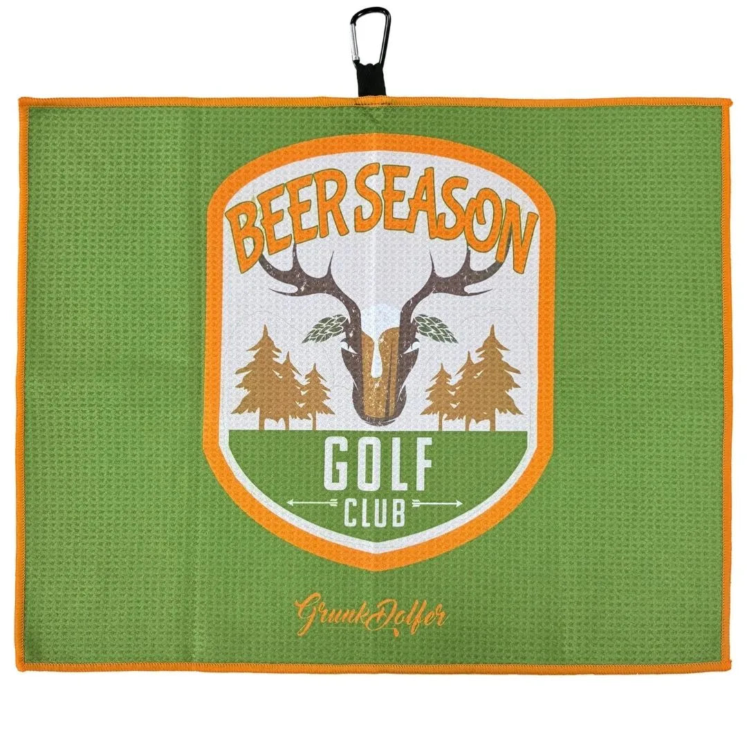 The Grunk Dolfer Beer Season Towel is green with an orange border and a badge reading "Beer Season Golf Club" featuring antlers, trees, and Grunkholfer at the bottom. It includes a black hanging loop at the top.