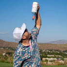 A man in a floral shirt and white hat tips an empty bottle of Island Boy by Grunk Dolfer into his open mouth on a golf course, with hills and a town in the background under a clear blue sky.