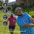 Two men golf outside on a lush course. Up front, one swings a club in the Grunk Dolfer Hibiscus Hitter blue and pink floral shirt, sunglasses, and beige cap. Icons show features; text: Matt is 6'3", 265 lbs, wearing XXL.