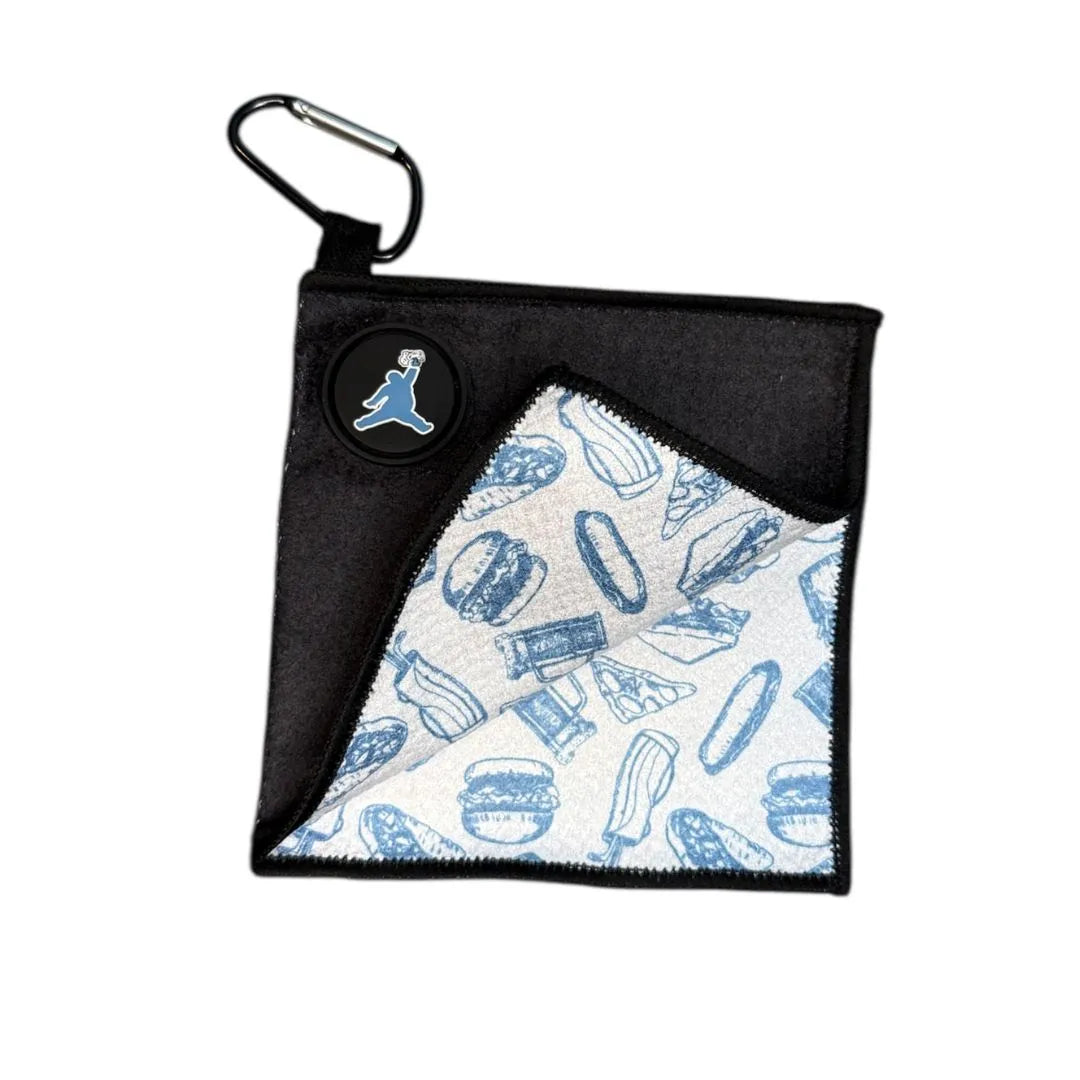 The Chunkman Golf Ball Towel by Grunk Dolfer is a black square microfiber towel with a logo patch and carabiner; a folded corner reveals a white inner layer featuring blue sketches of burgers, fries, hot dogs, and ice cream.